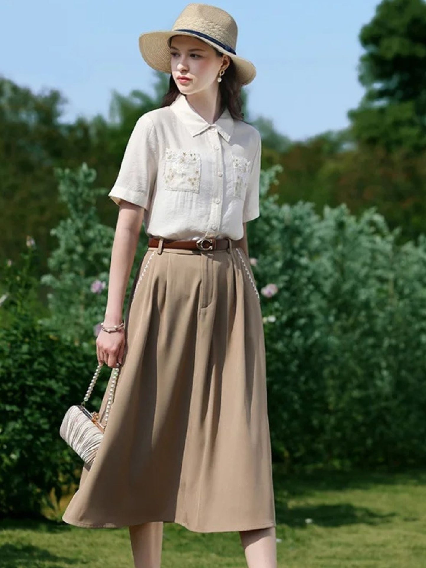 A woman wearing a white short-sleeved blouse with floral embroidery on the front pockets, a classic collar, and a button-down closure. She is also wearing a beige skirt, a brown belt, and a wide-brimmed hat.