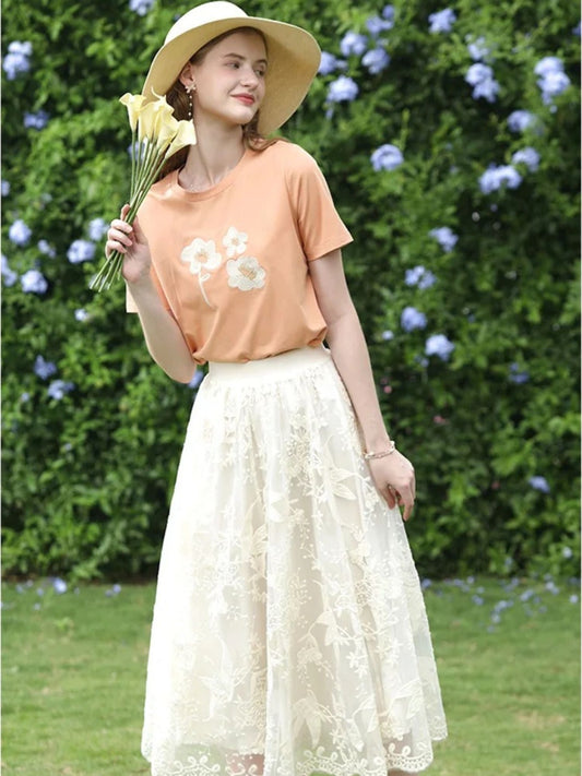 A woman wearing a beige embroidered lace midi skirt with a floral patterned skirt and an orange top, accessorized with a wide-brimmed hat.