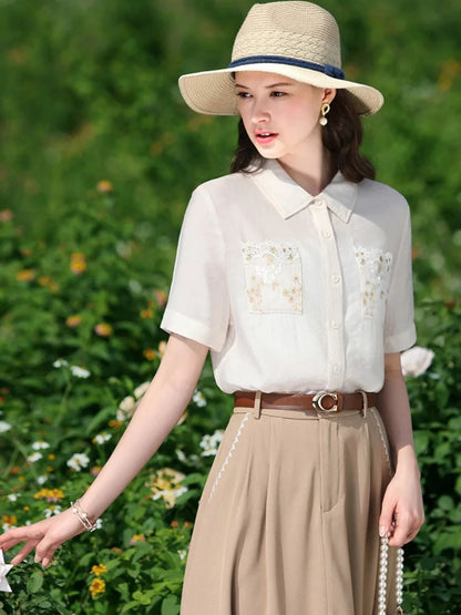A woman wearing a white short-sleeved blouse with floral embroidery on the front pockets, a classic collar, and a button-down closure. She is also wearing a beige skirt, a brown belt, and a wide-brimmed hat.