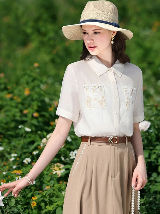 A woman wearing a white short-sleeved blouse with floral embroidery on the front pockets, a classic collar, and a button-down closure. She is also wearing a beige skirt, a brown belt, and a wide-brimmed hat.