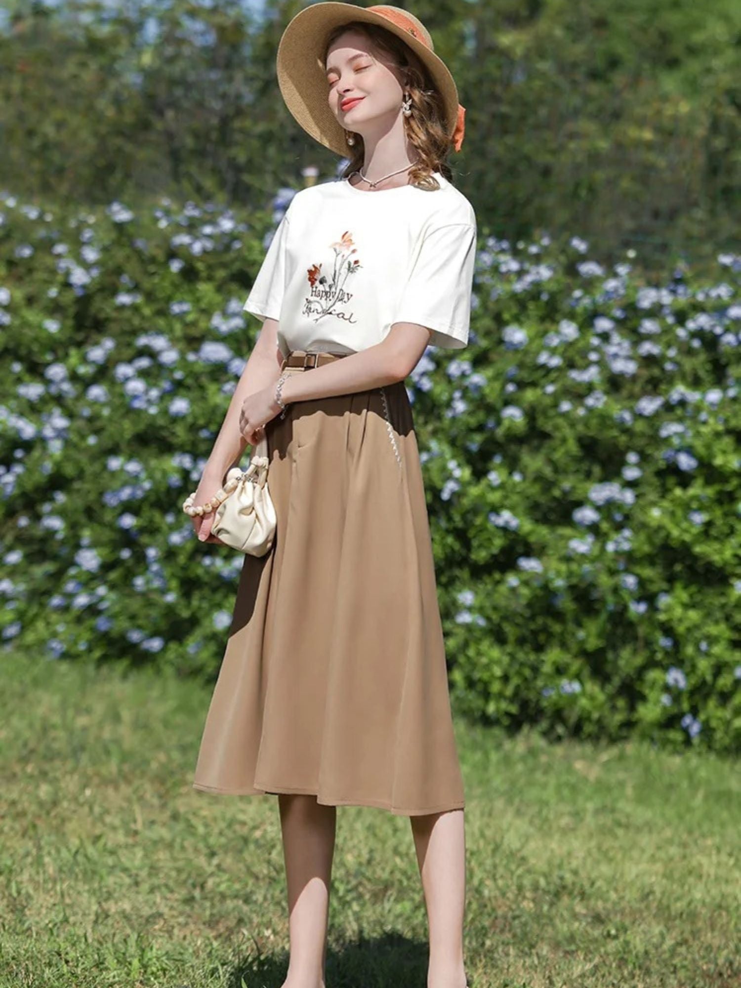 A woman wearing a high-waisted, midi-length beige skirt with an A-line silhouette, paired with a white t-shirt featuring a floral illustration, and accessorized with a wide-brimmed hat and a small handbag.