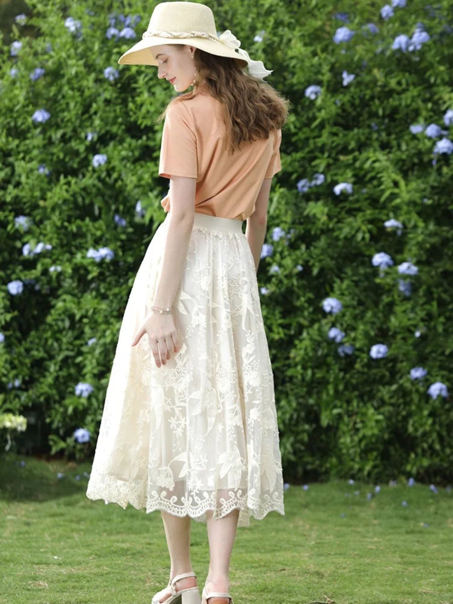 A woman wearing a beige embroidered lace midi skirt with a floral patterned skirt and an orange top, accessorized with a wide-brimmed hat.