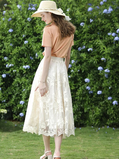 A woman wearing a beige embroidered lace midi skirt with a floral patterned skirt and an orange top, accessorized with a wide-brimmed hat.
