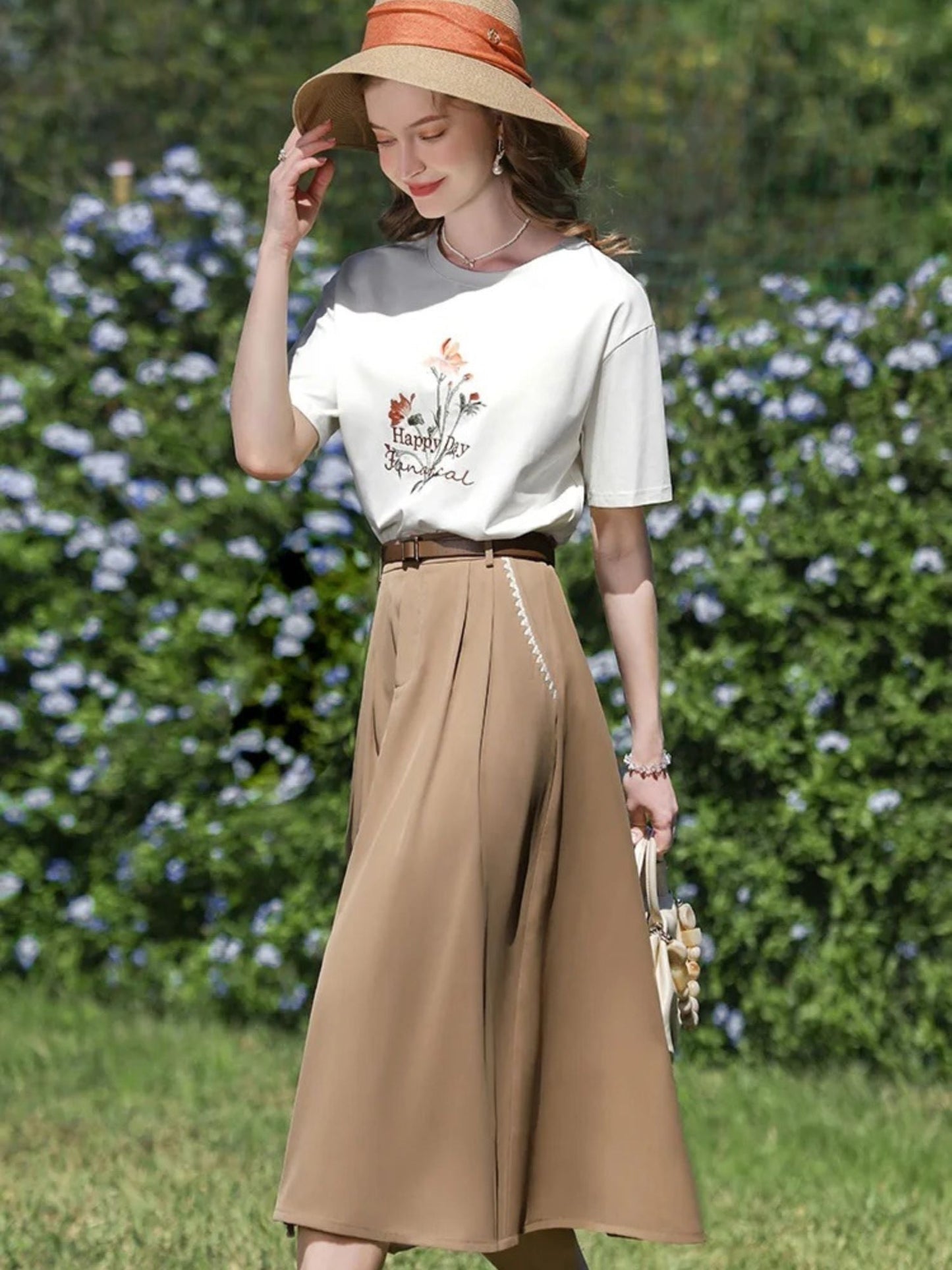 A woman wearing a high-waisted, midi-length beige skirt with an A-line silhouette, paired with a white t-shirt featuring a floral illustration, and accessorized with a wide-brimmed hat and a small handbag.
