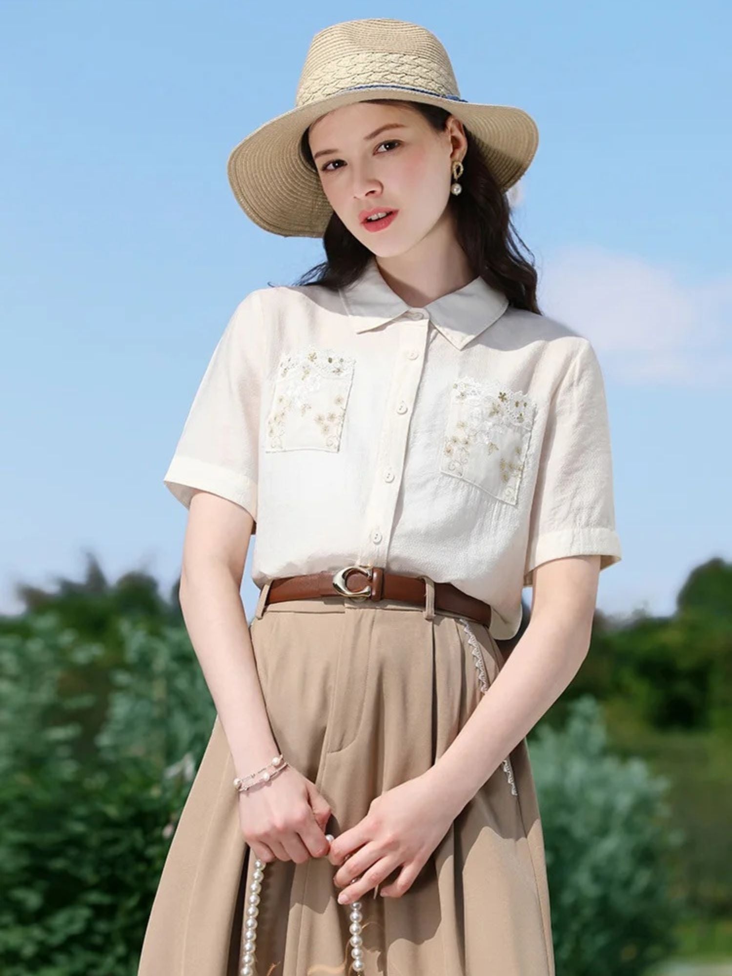 A woman wearing a white short-sleeved blouse with floral embroidery on the front pockets, a classic collar, and a button-down closure. She is also wearing a beige skirt, a brown belt, and a wide-brimmed hat.