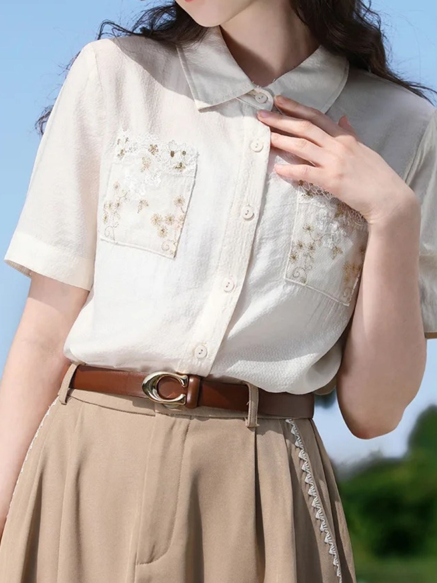 A woman wearing a white short-sleeved blouse with floral embroidery on the front pockets, a classic collar, and a button-down closure. She is also wearing a beige skirt, a brown belt, and a wide-brimmed hat.