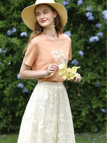 A woman wearing a beige embroidered lace midi skirt with a floral patterned skirt and an orange top, accessorized with a wide-brimmed hat.