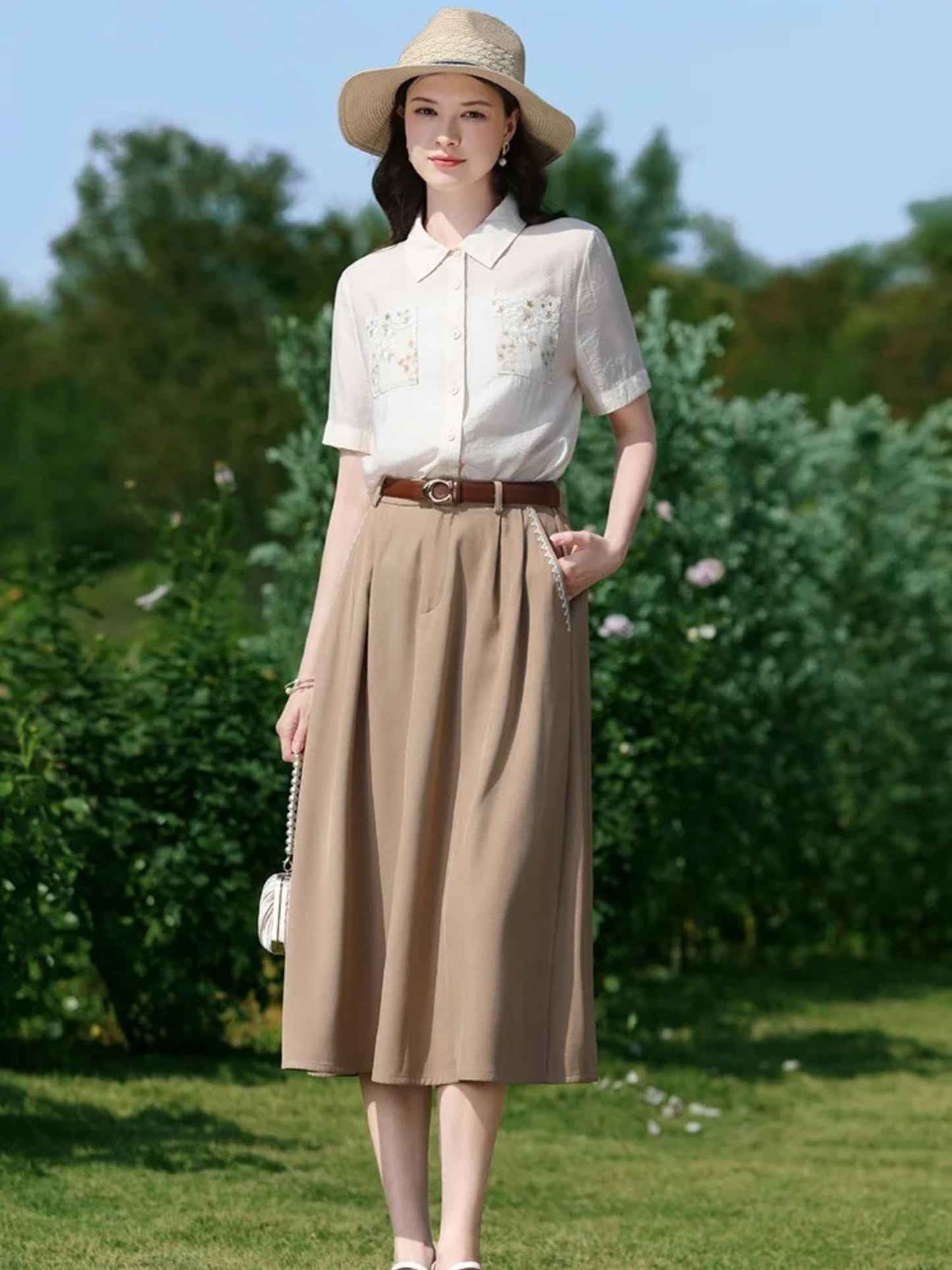 A woman wearing a white short-sleeved blouse with floral embroidery on the front pockets, a classic collar, and a button-down closure. She is also wearing a beige skirt, a brown belt, and a wide-brimmed hat.