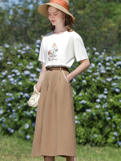 A woman wearing a high-waisted, midi-length beige skirt with an A-line silhouette, paired with a white t-shirt featuring a floral illustration, and accessorized with a wide-brimmed hat and a small handbag.