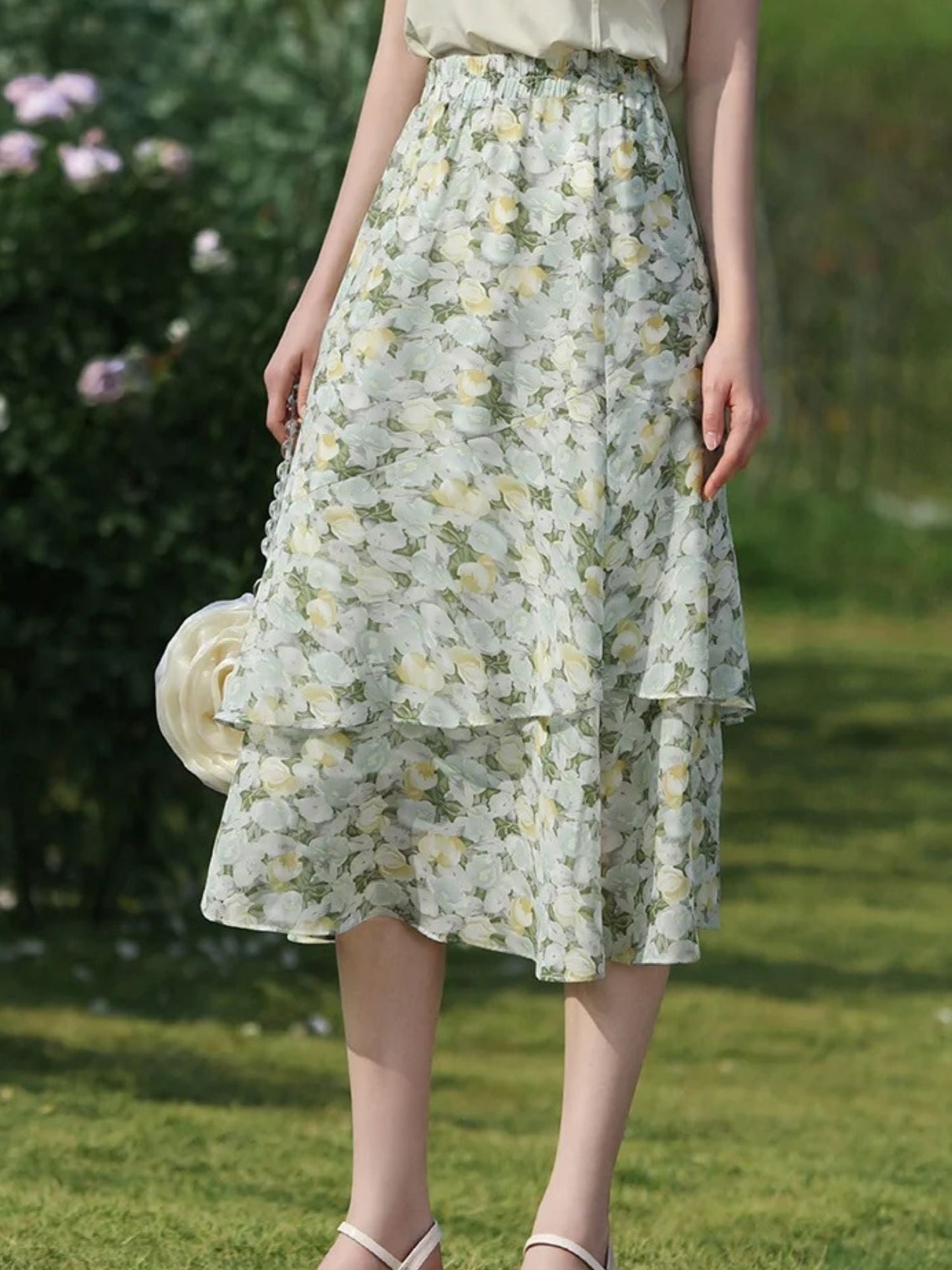 A woman wearing a floral chiffon midi skirt with a high elastic waistband, paired with a plain top, standing in a garden.
