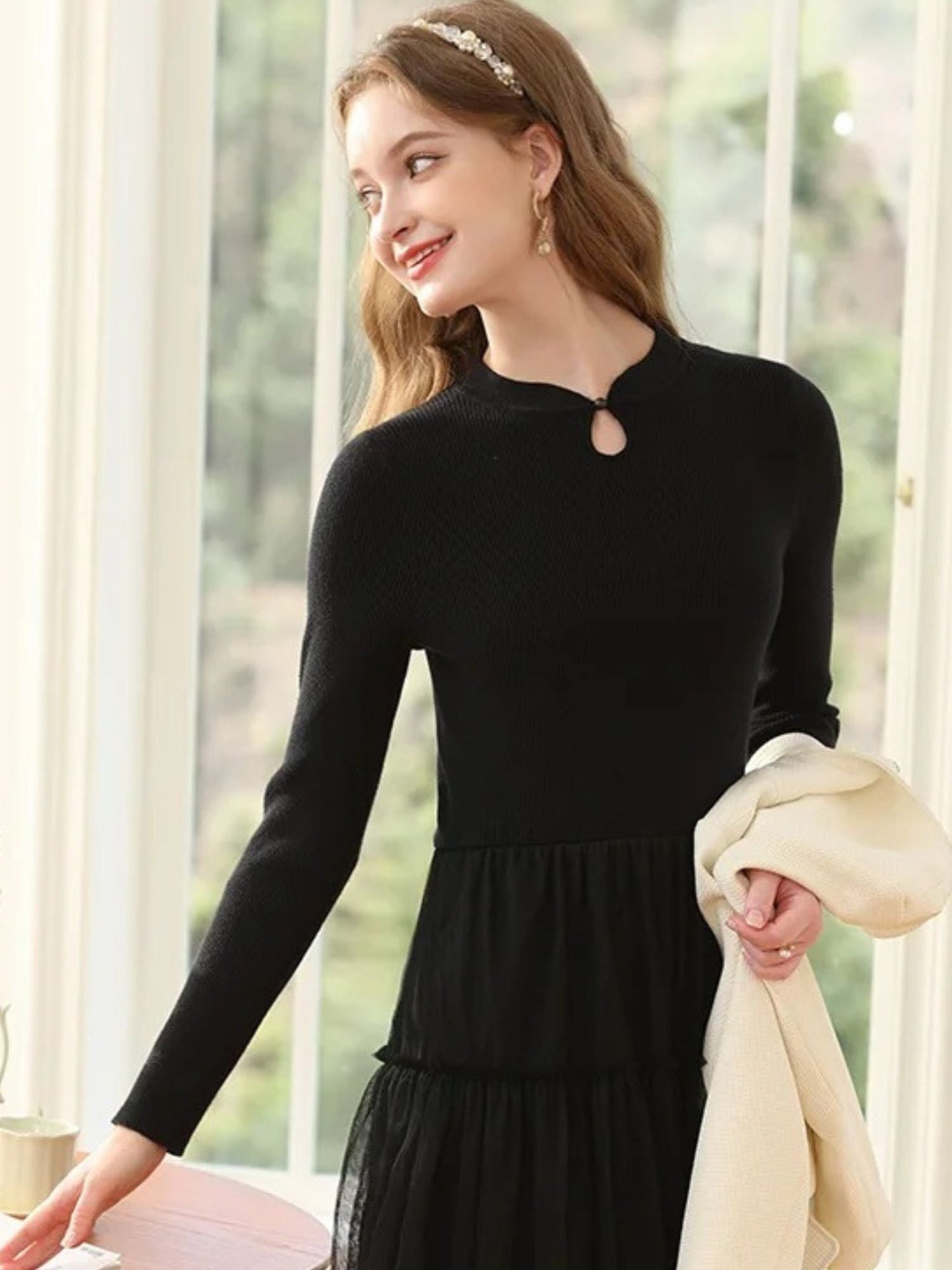 A woman wearing a long-sleeved, knee-length black dress with a tiered tulle skirt and a round neckline with a keyhole detail.