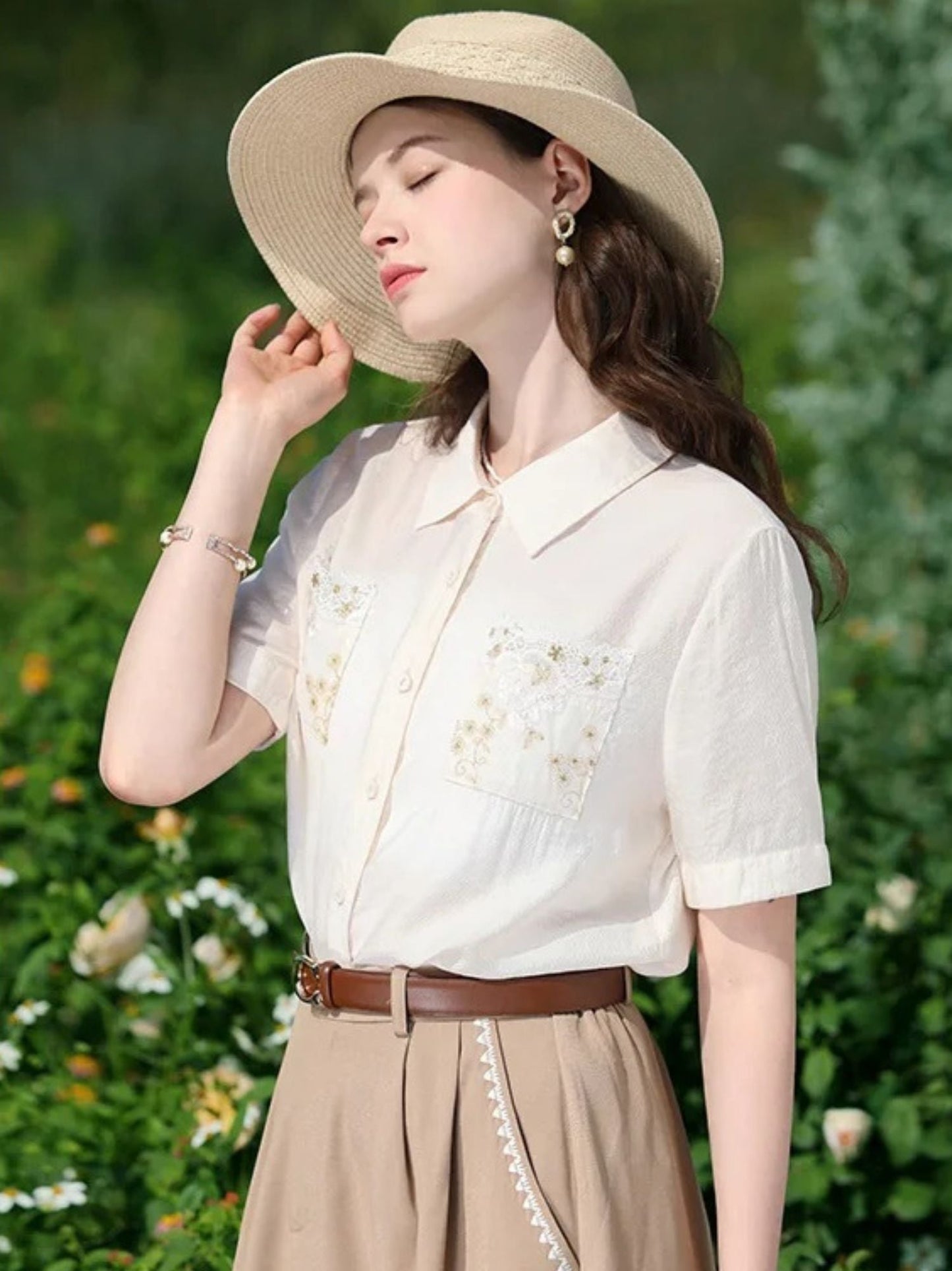 A woman wearing a white short-sleeved blouse with floral embroidery on the front pockets, a classic collar, and a button-down closure. She is also wearing a beige skirt, a brown belt, and a wide-brimmed hat.