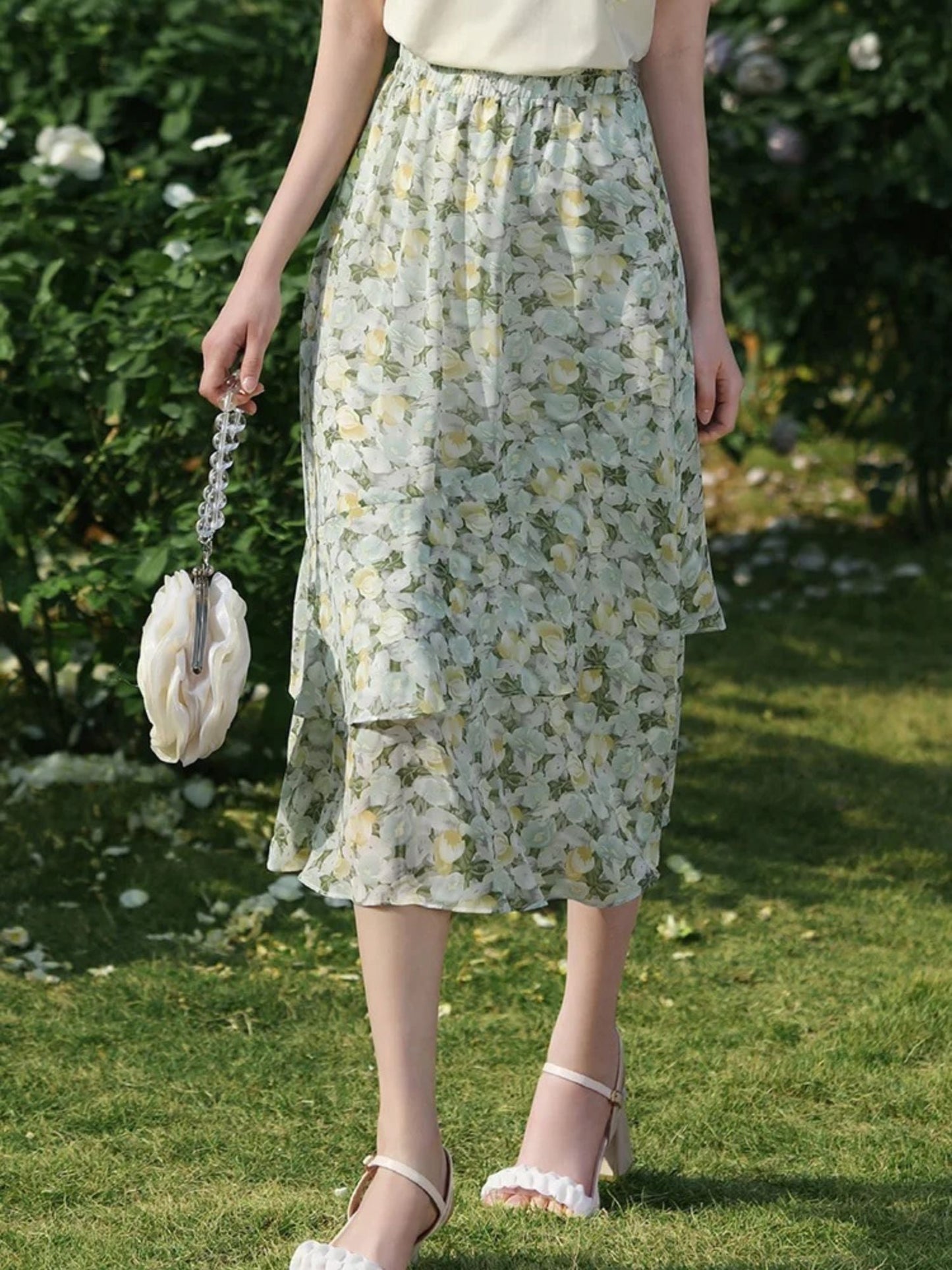 A woman wearing a floral chiffon midi skirt with a high elastic waistband, paired with a plain top, standing in a garden.