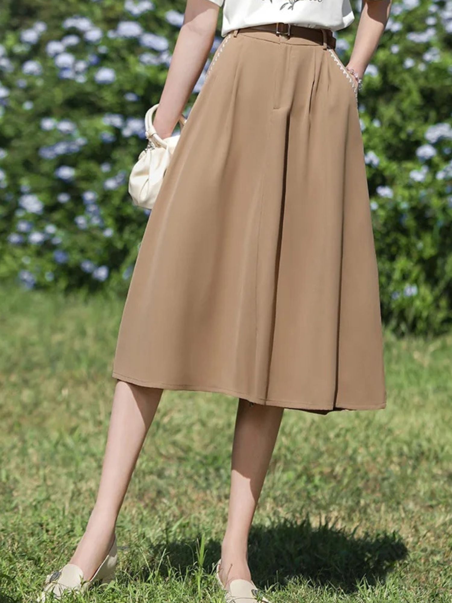A woman wearing a high-waisted, midi-length beige skirt with an A-line silhouette, paired with a white t-shirt featuring a floral illustration, and accessorized with a wide-brimmed hat and a small handbag.