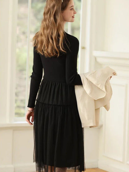 A woman wearing a long-sleeved, knee-length black dress with a tiered tulle skirt and a round neckline with a keyhole detail.