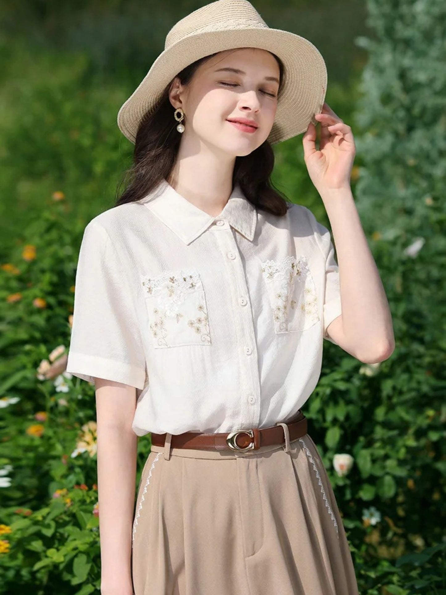 A woman wearing a white short-sleeved blouse with floral embroidery on the front pockets, a classic collar, and a button-down closure. She is also wearing a beige skirt, a brown belt, and a wide-brimmed hat.